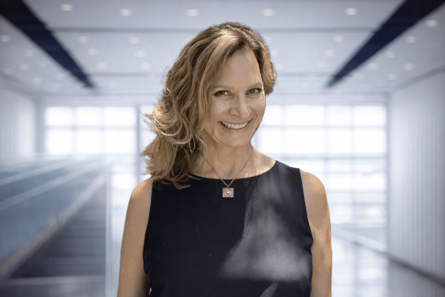 Woman with wavy hair wearing a sleeveless black top and a necklace, smiling, standing in a bright modern indoor setting with large windows.