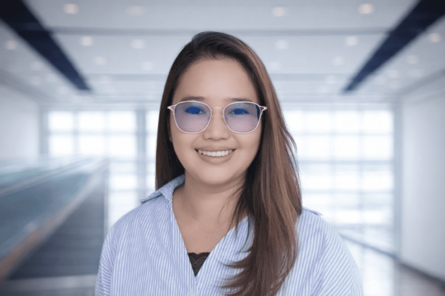 A woman with long brown hair and blue glasses smiles at the camera, standing in a modern, bright indoor setting with large windows.