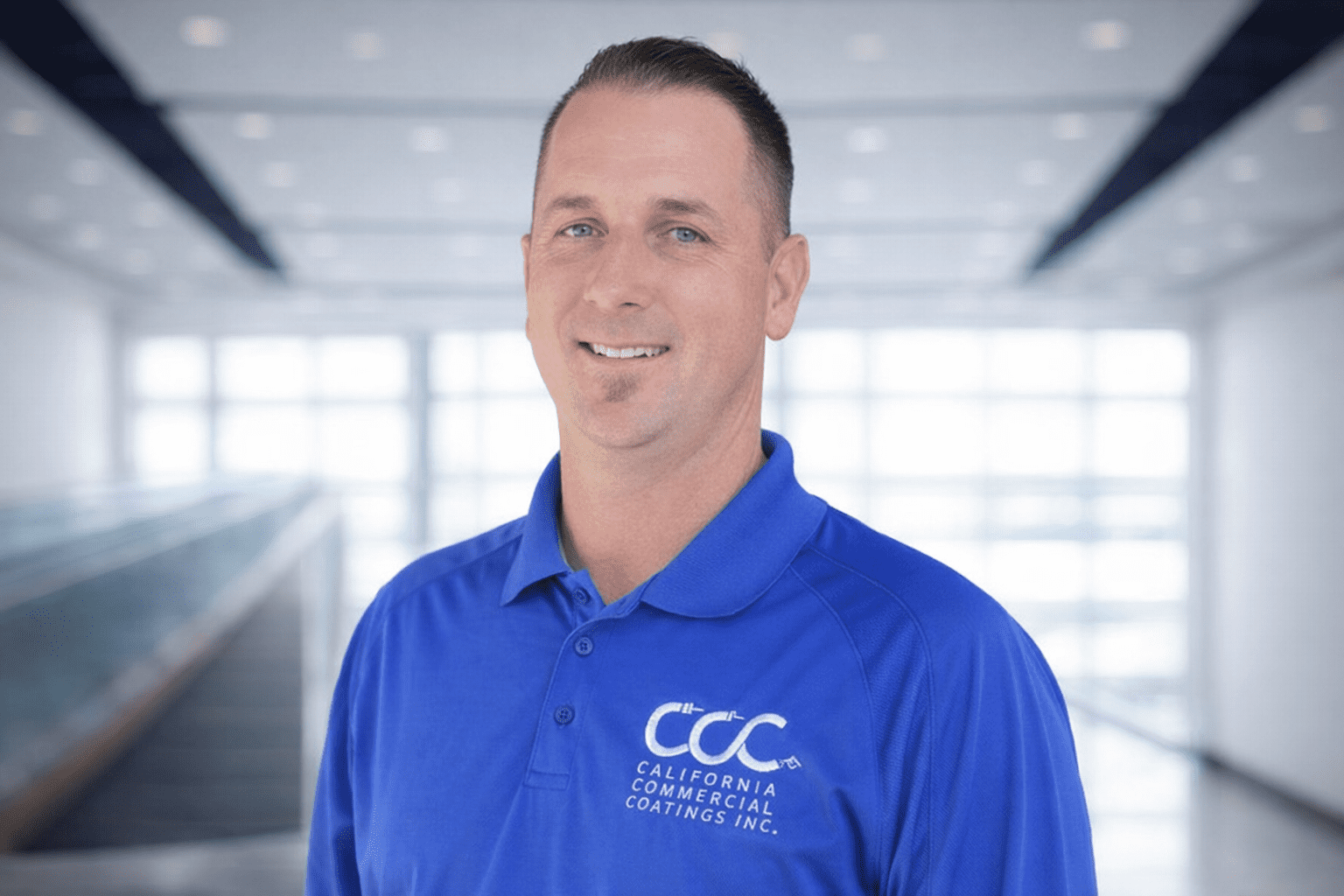 A man in a blue "California Commercial Coatings Inc." polo shirt stands indoors, smiling at the camera with a blurred modern background.