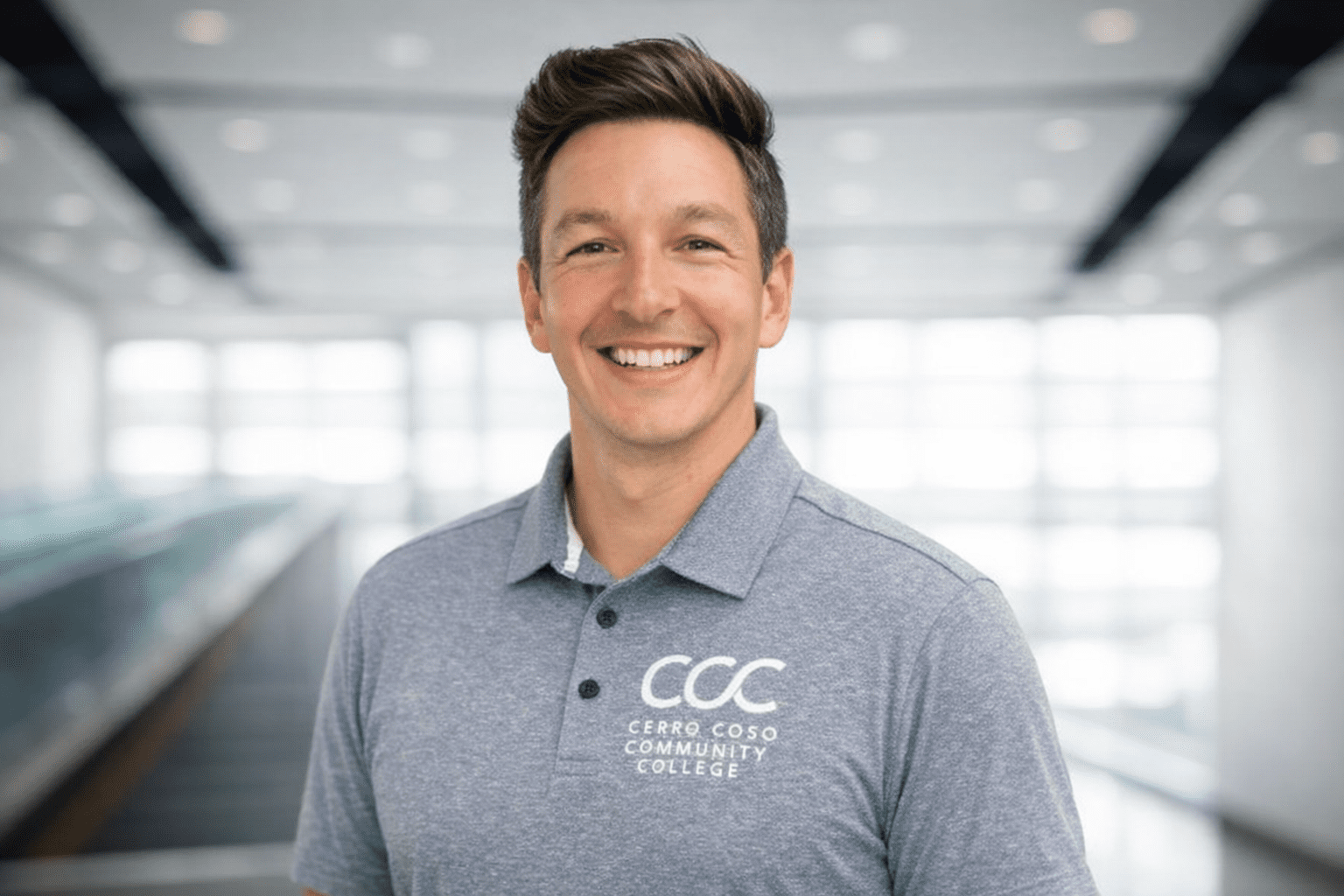 A smiling person wearing a light blue Cerro Coso Community College polo shirt stands indoors with a blurred, bright background.