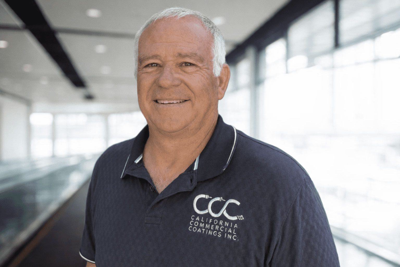 A man with short gray hair wearing a navy blue "California Commercial Coatings Inc." polo shirt stands indoors near large windows.